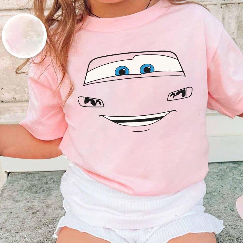 Op de afbeelding: Een lichtroze T-shirt met korte mouwen en een grafische print van een autogezicht met grote blauwe ogen, zwarte omlijningen en een brede glimlachende mond. Het shirt wordt gecombineerd met een witte short met ruches.