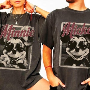 May include: Two charcoal gray t-shirts featuring Mickey and Minnie Mouse. Each shirt has a framed graphic of the character wearing sunglasses, with the character's name in a burgundy script above the image. The shirts are a relaxed fit.