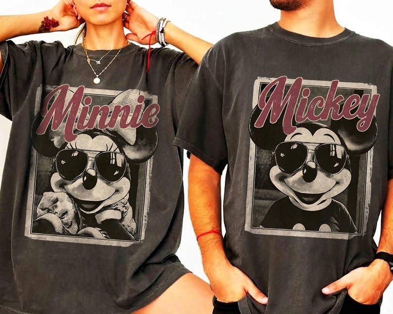 May include: Two charcoal gray t-shirts featuring Mickey and Minnie Mouse. Each shirt has a framed graphic of the character wearing sunglasses, with the character's name in a burgundy script above the image. The shirts are a relaxed fit.