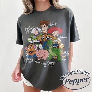 May include: Dark grey t-shirt with a graphic of Toy Story characters, including Woody, Buzz Lightyear, and Jessie. The text "Toy Story" is written in a script font. The shirt also has the text "Comfort Colors Pepper".