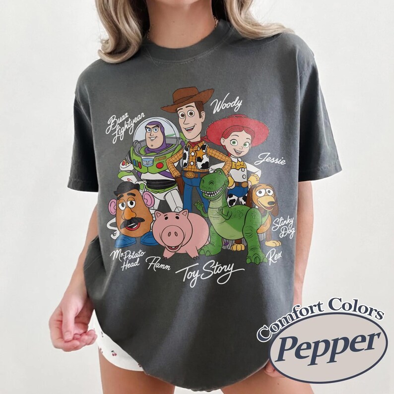 May include: Dark grey t-shirt with a graphic of Toy Story characters, including Woody, Buzz Lightyear, and Jessie. The text "Toy Story" is written in a script font. The shirt also has the text "Comfort Colors Pepper".