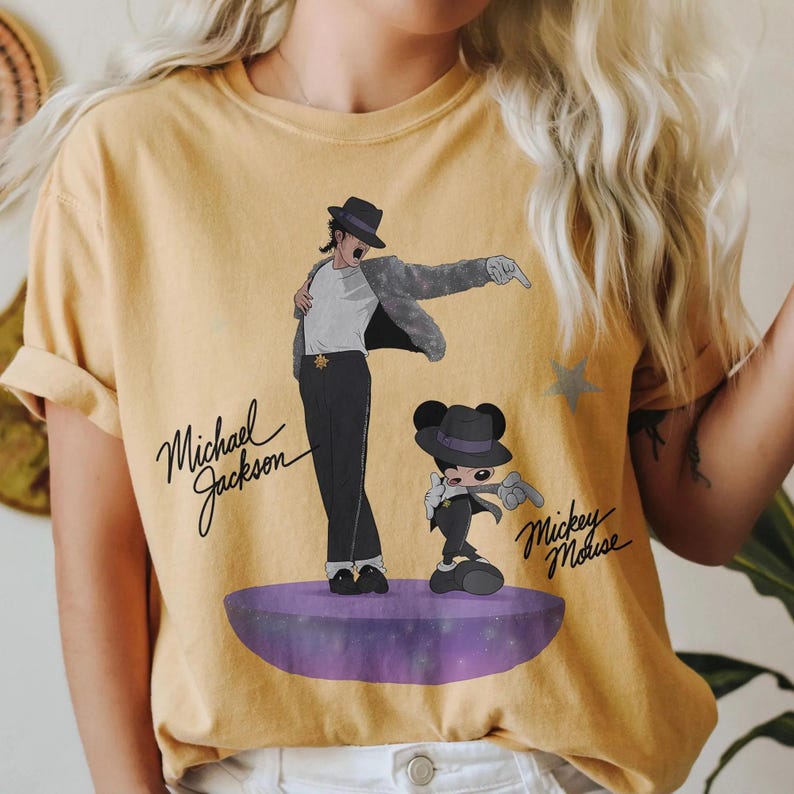 Puede incluir: Camiseta de color amarillo mostaza con un gr&aacute;fico de Michael Jackson y Mickey Mouse con trajes a juego. Los textos "Michael Jackson" y "Mickey Mouse" est&aacute;n escritos en cursiva. Las figuras est&aacute;n sobre un escenario morado.