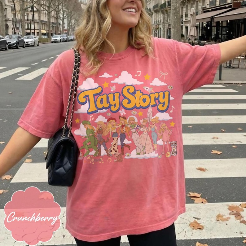 May include: A coral-colored short-sleeve t-shirt featuring the text Tay Story in yellow with a blue outline. The graphic includes a group of illustrated characters, pink clouds, stars, and musical notes. A black quilted shoulder bag is also visible.