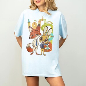 May include: Pale blue t-shirt with a cartoon image of a fox and a rabbit. The fox is in a suit, the rabbit in a police uniform. The graphic has a colourful, whimsical design, with a background of stylized shapes.