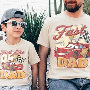 May include: Two beige t-shirts with matching graphics. The shirts feature a red race car with the text "Fast Like 95 Dad". One shirt is worn by a child wearing sunglasses and a cap. The other shirt is worn by an adult.