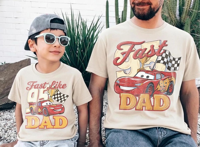 May include: Two beige t-shirts with matching graphics. The shirts feature a red race car with the text "Fast Like 95 Dad". One shirt is worn by a child wearing sunglasses and a cap. The other shirt is worn by an adult.
