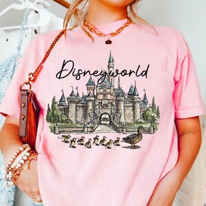 Puede incluir: Camiseta rosa claro con un dise&ntilde;o de Disneyworld. El dise&ntilde;o incluye un castillo, patos y la palabra "Disneyworld" en escritura cursiva. La camiseta es de color rosa claro.