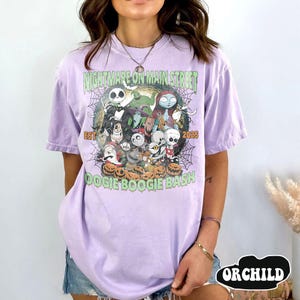 Puede incluir: Una camiseta de color lila claro con un dise&ntilde;o gr&aacute;fico que presenta personajes de una pel&iacute;cula. El dise&ntilde;o incluye el texto "NIGHTMARE ON MAIN STREET" y "DOOGIE BOOGIE BASH" con el a&ntilde;o "2025". La camiseta tiene mangas cortas.