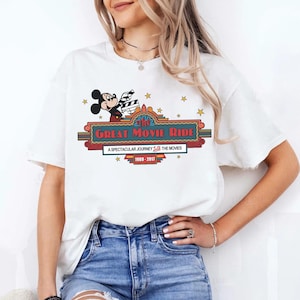 Puede incluir: Camiseta blanca con Mickey Mouse sosteniendo una claqueta de cine sobre el texto "The Great Movie Ride". El dise&ntilde;o incluye estrellas y el texto "A Spectacular Journey Through The Movies 1989-2017".