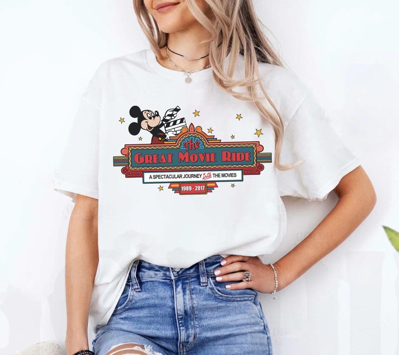 Puede incluir: Camiseta blanca con Mickey Mouse sosteniendo una claqueta de cine sobre el texto "The Great Movie Ride". El dise&ntilde;o incluye estrellas y el texto "A Spectacular Journey Through The Movies 1989-2017".