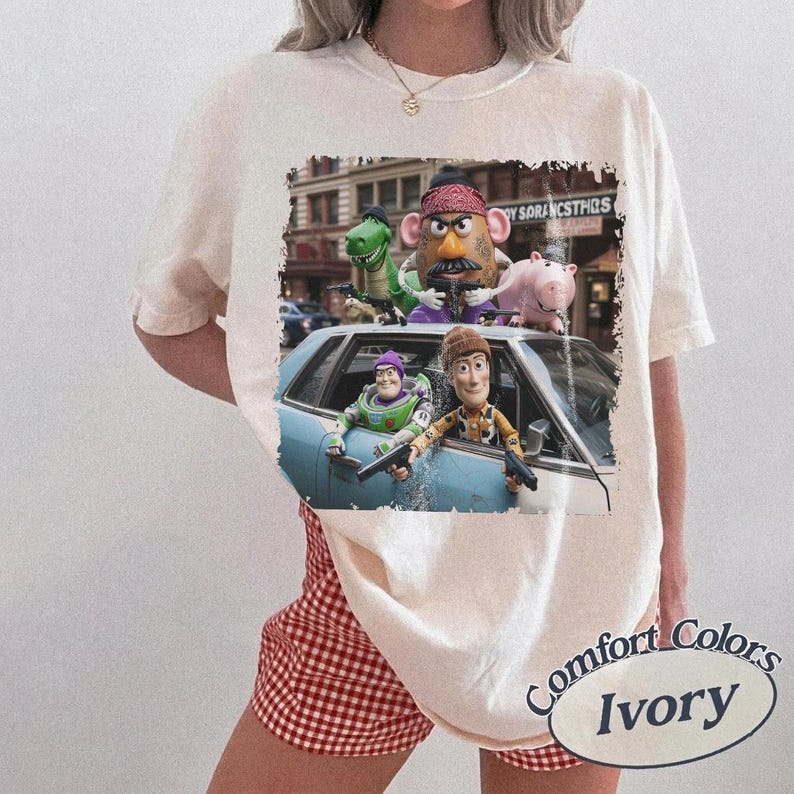 K&ouml;nnte beinhalten: Ein elfenbeinfarbenes T-Shirt mit einem Grafikdruck der Toy Story-Figuren, darunter Woody, Buzz Lightyear, Mr. Potato Head, Rex und Hamm. Die Figuren sind in und um ein hellblaues Auto vor einer st&auml;dtischen Kulisse dargestellt.