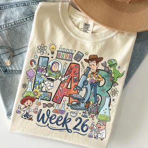 May include: Cream-colored t-shirt featuring a colorful graphic with the text "LAB Week 26." The design includes cartoon characters and lab equipment. The shirt is folded on top of blue jeans.