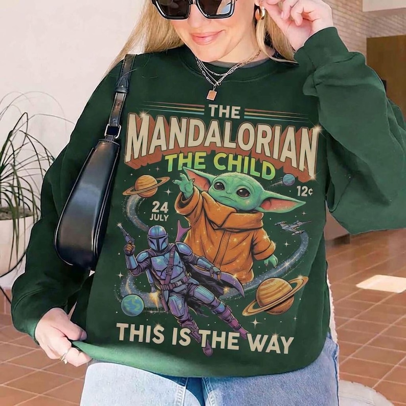 May include: Dark green sweatshirt featuring a graphic design from The Mandalorian series. The design includes the text "The Mandalorian", "The Child", and "This is the Way", along with images of Baby Yoda and the Mandalorian.