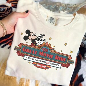 Puede incluir: Sudadera de color crema con Mickey Mouse sosteniendo una claqueta de cine. El dise&ntilde;o incluye el texto "The Great Movie Ride" en rojo y azul, con los a&ntilde;os 1989-2017. Estrellas doradas salpican el gr&aacute;fico.