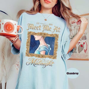 May include: Light blue t-shirt with the text "Meet Me At Midnight" and a framed watercolor illustration of a woman. The shirt also features the text "Est. 1980". A white and orange mug with a sun design is held in the image.