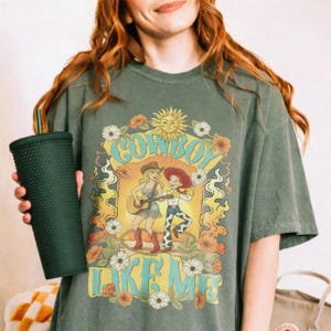May include: A dark olive green t-shirt featuring a graphic print of two cartoon characters, one playing a guitar, surrounded by flowers and a sun. The text reads COWBOY LIKE ME. A dark green studded tumbler with a straw is held in the foreground.