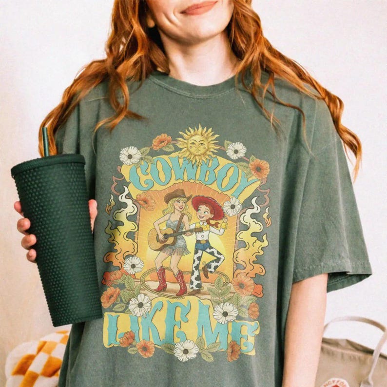 May include: A dark olive green t-shirt featuring a graphic print of two cartoon characters, one playing a guitar, surrounded by flowers and a sun. The text reads COWBOY LIKE ME. A dark green studded tumbler with a straw is held in the foreground.