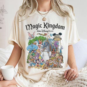 Puede incluir: Camiseta color crema con un gr&aacute;fico colorido de varios personajes de Disney, incluyendo Mickey Mouse, Aladdin y Blancanieves. La camiseta tiene las palabras "Magic Kingdom" y "Walt Disney World" impresas en negro.