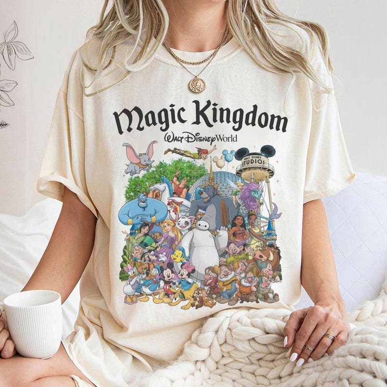 Puede incluir: Camiseta color crema con un gr&aacute;fico colorido de varios personajes de Disney, incluyendo Mickey Mouse, Aladdin y Blancanieves. La camiseta tiene las palabras "Magic Kingdom" y "Walt Disney World" impresas en negro.