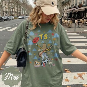May include: An olive green t-shirt featuring a graphic print of a person with long hair playing a guitar, wearing a yellow plaid shirt and cow-print pants. The design includes blue clouds, flowers, a red hat, and the letters TS in yellow.