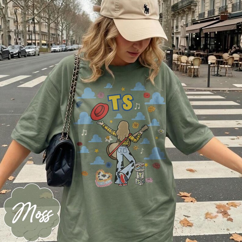 May include: An olive green t-shirt featuring a graphic print of a person with long hair playing a guitar, wearing a yellow plaid shirt and cow-print pants. The design includes blue clouds, flowers, a red hat, and the letters TS in yellow.