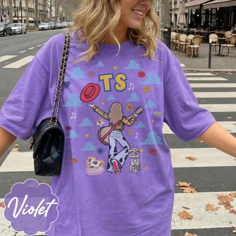 May include: A purple t-shirt featuring a graphic illustration of a person with long hair playing a guitar, wearing a yellow plaid shirt and cow-print pants. The design includes the text TS, a red hat, blue clouds, flowers, and stacked blocks.