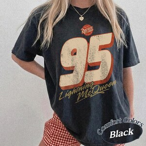 Puede incluir: Camiseta negra con un dise&ntilde;o desgastado que muestra el n&uacute;mero "95" en crema y rojo, y el texto "Lightning McQueen". La camiseta tambi&eacute;n tiene el texto "Rusteze" y "Comfort Colors Black".