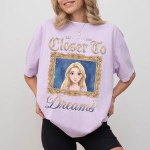 May include: A light purple t-shirt featuring a framed image of a cartoon character with long blonde hair. The text "Closer To Dreams" and "Est. 2010" are printed on the shirt in gold and blue script.