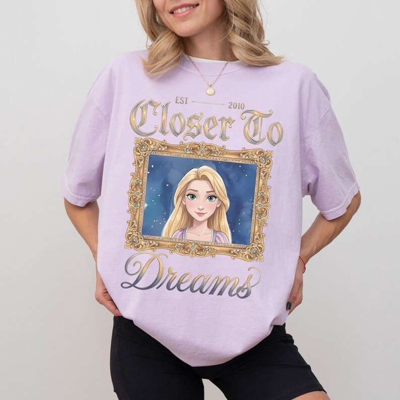 May include: A light purple t-shirt featuring a framed image of a cartoon character with long blonde hair. The text "Closer To Dreams" and "Est. 2010" are printed on the shirt in gold and blue script.