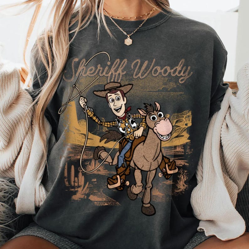 K&ouml;nnte beinhalten: Dunkelgraues Sweatshirt mit Sheriff Woody aus Toy Story auf einem Pferd, mit dem Text "Sheriff Woody" &uuml;ber der Grafik. Das Design hat einen Vintage-Look mit einem W&uuml;stenlandschaftshintergrund.
