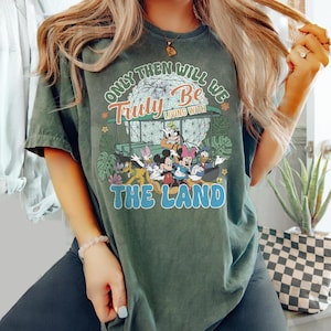 Könnte beinhalten: Dunkelgrünes T-Shirt mit einem farbenfrohen Grafikdesign von Disney-Figuren und dem Text "Only Then Will We Truly Be Living With The Land." Das Design zeigt Micky Maus, Minnie Maus, Donald Duck und Goofy. Das Shirt hat einen Vintage-Look.