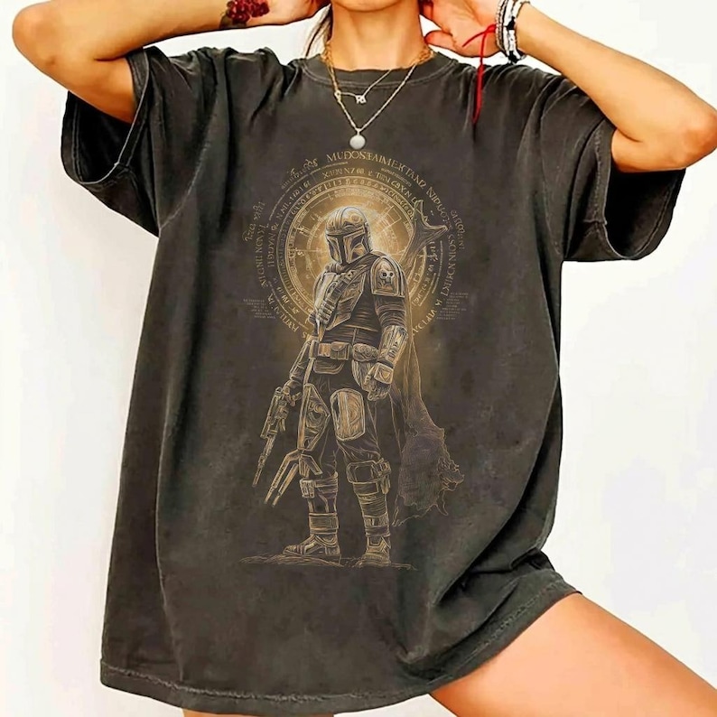 May include: Dark grey t-shirt with a detailed illustration of a Mandalorian warrior. The design includes a circular emblem with text and a depiction of the character in full armour, holding a weapon. The shirt has a vintage, worn look.