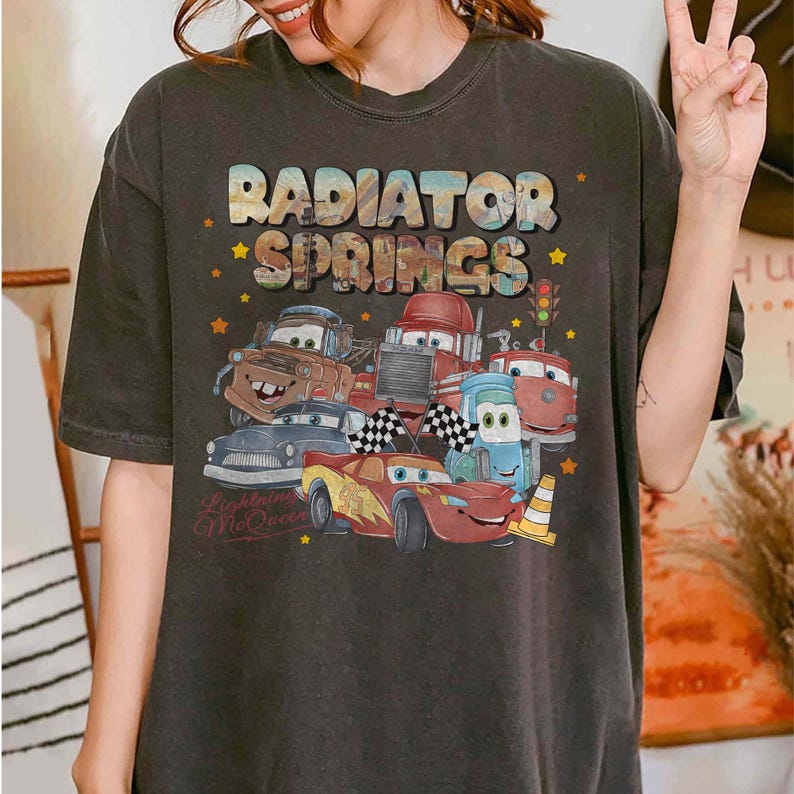May include: Dark gray t-shirt featuring a colorful graphic of characters from the movie Cars, including Lightning McQueen. The text "RADIATOR SPRINGS" is displayed above the characters. The shirt has a vintage, worn look.