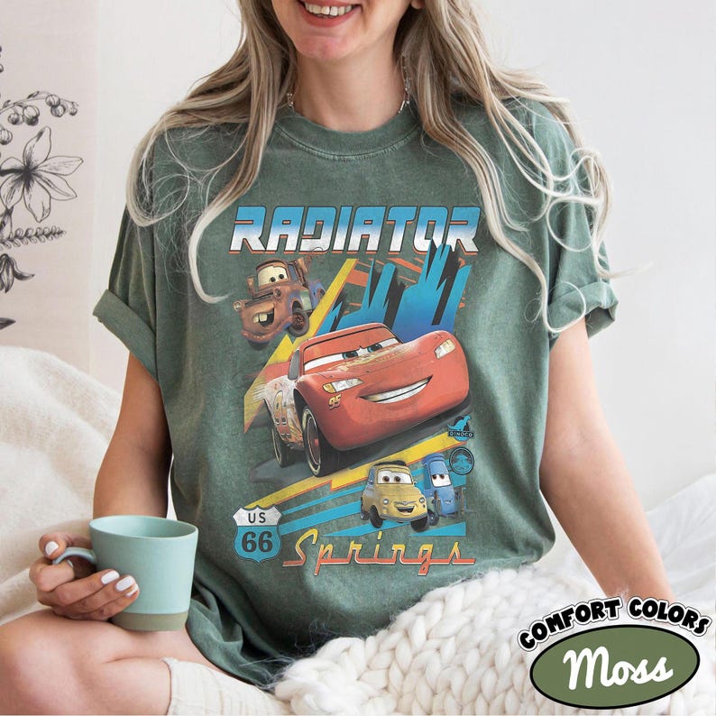 May include: A moss green short-sleeve t-shirt featuring a graphic print of characters from the movie Cars, including Lightning McQueen, Mater, and Luigi. The text reads Radiator Springs and US 66. The shirt has a crew neckline and a relaxed fit.