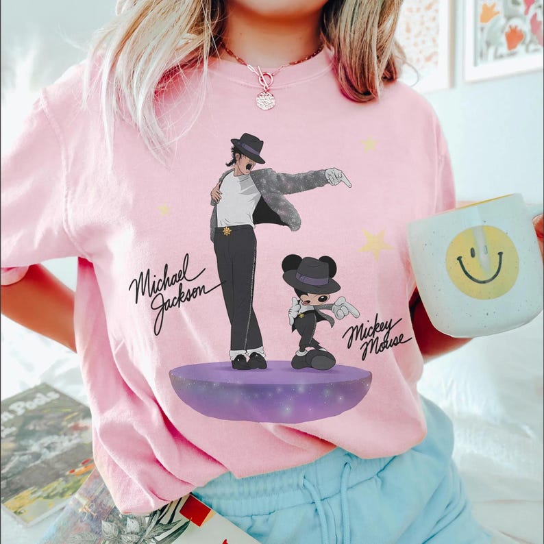 Puede incluir: Camiseta rosa claro con un dise&ntilde;o de Michael Jackson y Mickey Mouse. La ilustraci&oacute;n muestra a Michael Jackson en su pose ic&oacute;nica y a Mickey Mouse con un atuendo similar. Los nombres "Michael Jackson" y "Mickey Mouse" tambi&eacute;n est&aacute;n impresos.