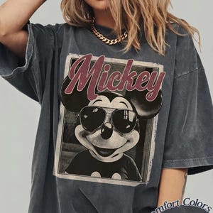 May include: Dark grey t-shirt featuring a graphic of Mickey Mouse wearing sunglasses, with the name "Mickey" in a burgundy script. The shirt has a vintage, faded look and the words "Comfort Colors Pepper" are printed on the bottom right.