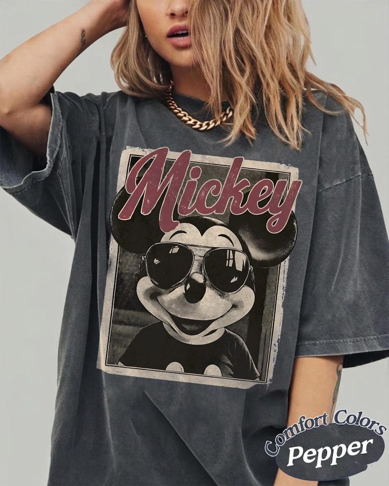 May include: Dark grey t-shirt featuring a graphic of Mickey Mouse wearing sunglasses, with the name "Mickey" in a burgundy script. The shirt has a vintage, faded look and the words "Comfort Colors Pepper" are printed on the bottom right.