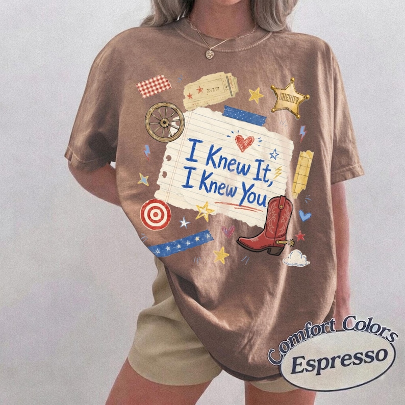 May include: An espresso-colored t-shirt featuring a graphic print of a lined paper scrap with the text I Knew It, I Knew You, a red cowboy boot, a sheriff star, a wagon wheel, a target, a ticket, and various small stars and blue tape accents.