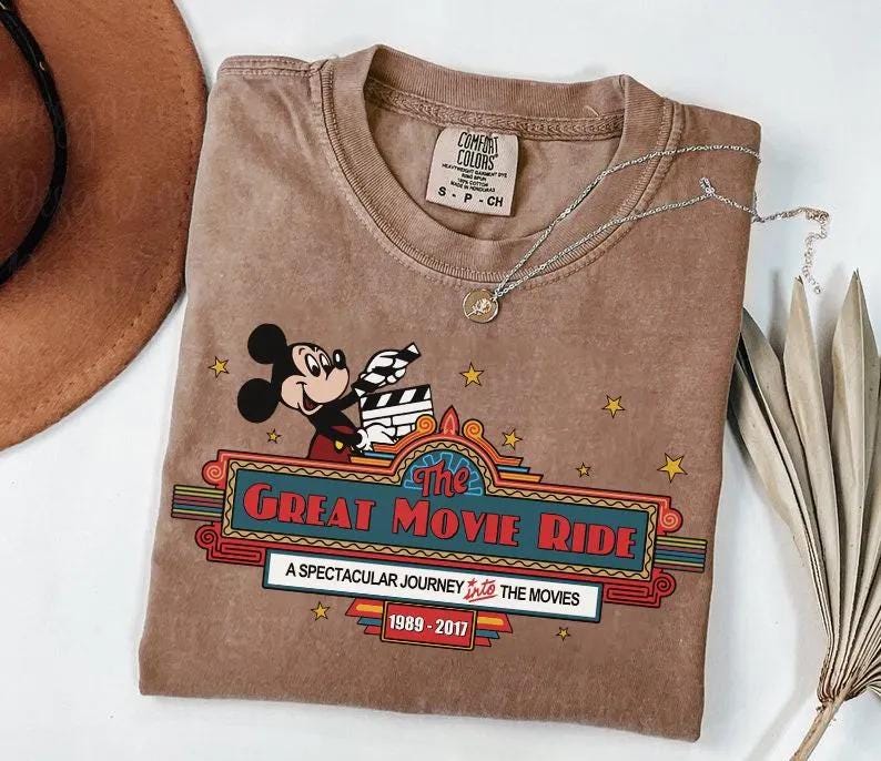 Puede incluir: Camiseta marr&oacute;n con Mickey Mouse sosteniendo una claqueta de cine, con el texto "The Great Movie Ride" y los a&ntilde;os 1989-2017. La camiseta tiene cuello redondo y una peque&ntilde;a etiqueta. Un collar plateado con un colgante est&aacute; sobre la camiseta.