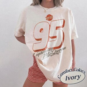 Puede incluir: Camiseta de color marfil con un gran n&uacute;mero 95 naranja y las palabras "Lightning McQueen" y "Rusteze". La camiseta tiene un aspecto vintage y desgastado. La esquina inferior derecha de la camiseta tiene las palabras "Comfort Colors" e "Ivory".