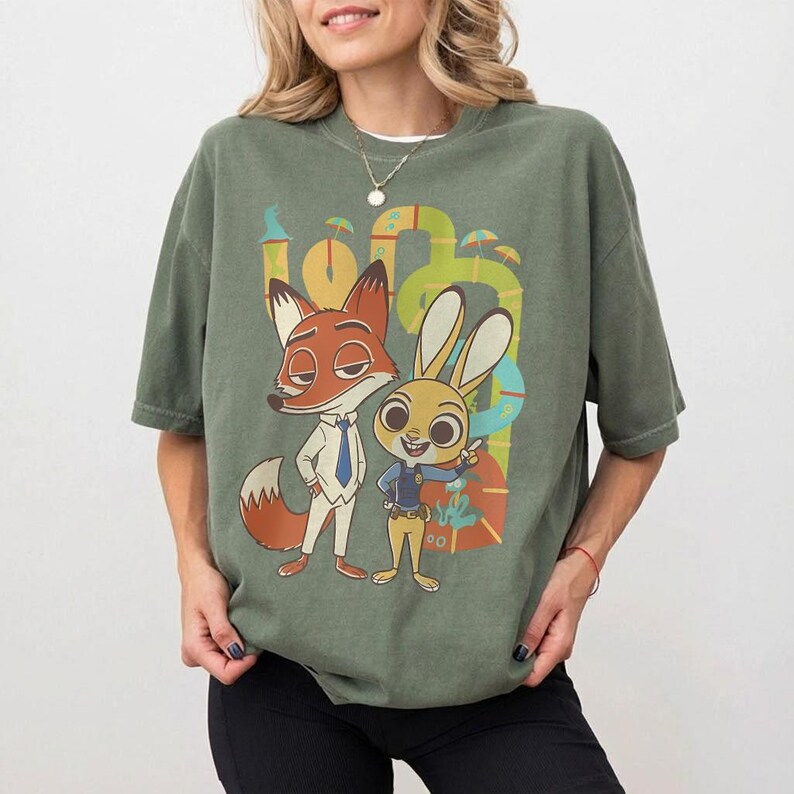 May include: Sage green t-shirt with a cartoon image of Nick Wilde and Judy Hopps from Zootropolis. Nick, a fox, wears a suit, and Judy, a rabbit, is in a police uniform. The background has a colourful, abstract design.