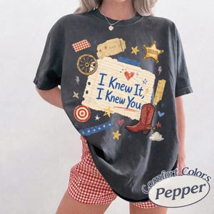 May include: A charcoal gray short-sleeve t-shirt featuring a graphic print of a lined notebook paper with the text I Knew It, I Knew You, surrounded by illustrations of a red cowboy boot, a sheriff star, a wagon wheel, and various small stars.