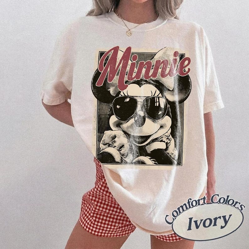 May include: Ivory-colored t-shirt featuring a black and white graphic of Minnie Mouse wearing sunglasses, with the name "Minnie" in a burgundy script. The shirt is part of the Comfort Colors collection.