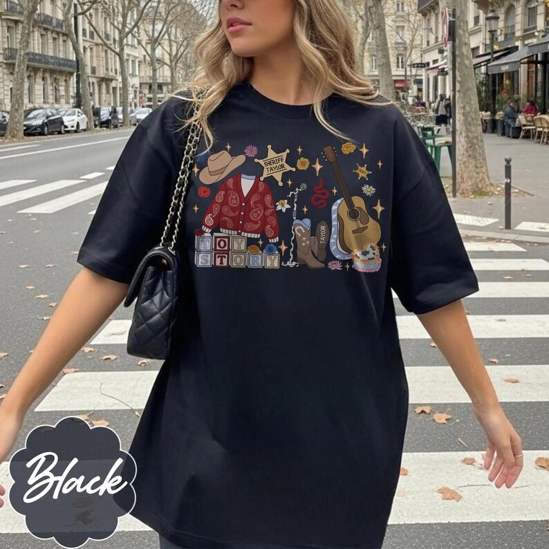 May include: A black t-shirt featuring a graphic print of a brown cowboy hat, a red paisley cardigan, a sheriff badge labeled Taylor, a guitar, a cowboy boot, and blocks spelling Toy Story, accented with small stars and floral elements.