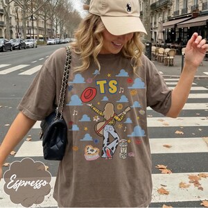 May include: A brown t-shirt featuring a graphic print of a person with long hair wearing a yellow plaid shirt and cow-print pants, holding a guitar. The design includes blue clouds, a red hat, the text TS, and blocks with the letters TS.
