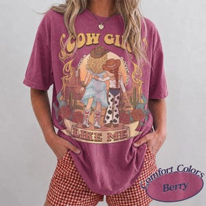 Puede incluir: Una camiseta de manga corta color baya con un estampado gr&aacute;fico de dos figuras con sombreros y botas de vaquero en un paisaje des&eacute;rtico con cactus y rosas. El texto dice COW GIRL LIKE ME. La prenda tiene un cuello redondo y corte holgado.