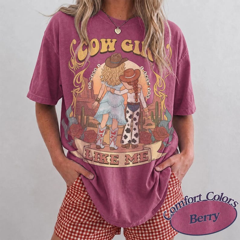 Puede incluir: Una camiseta de manga corta color baya con un estampado gr&aacute;fico de dos figuras con sombreros y botas de vaquero en un paisaje des&eacute;rtico con cactus y rosas. El texto dice COW GIRL LIKE ME. La prenda tiene un cuello redondo y corte holgado.