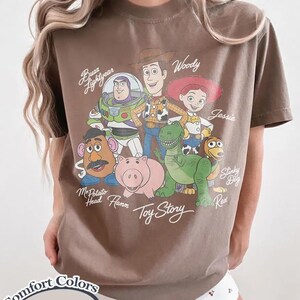 May include: Espresso-coloured t-shirt featuring a graphic of Toy Story characters including Buzz Lightyear, Woody, and Jessie. The shirt also has the text "Toy Story" and "Comfort Colors" printed on it.