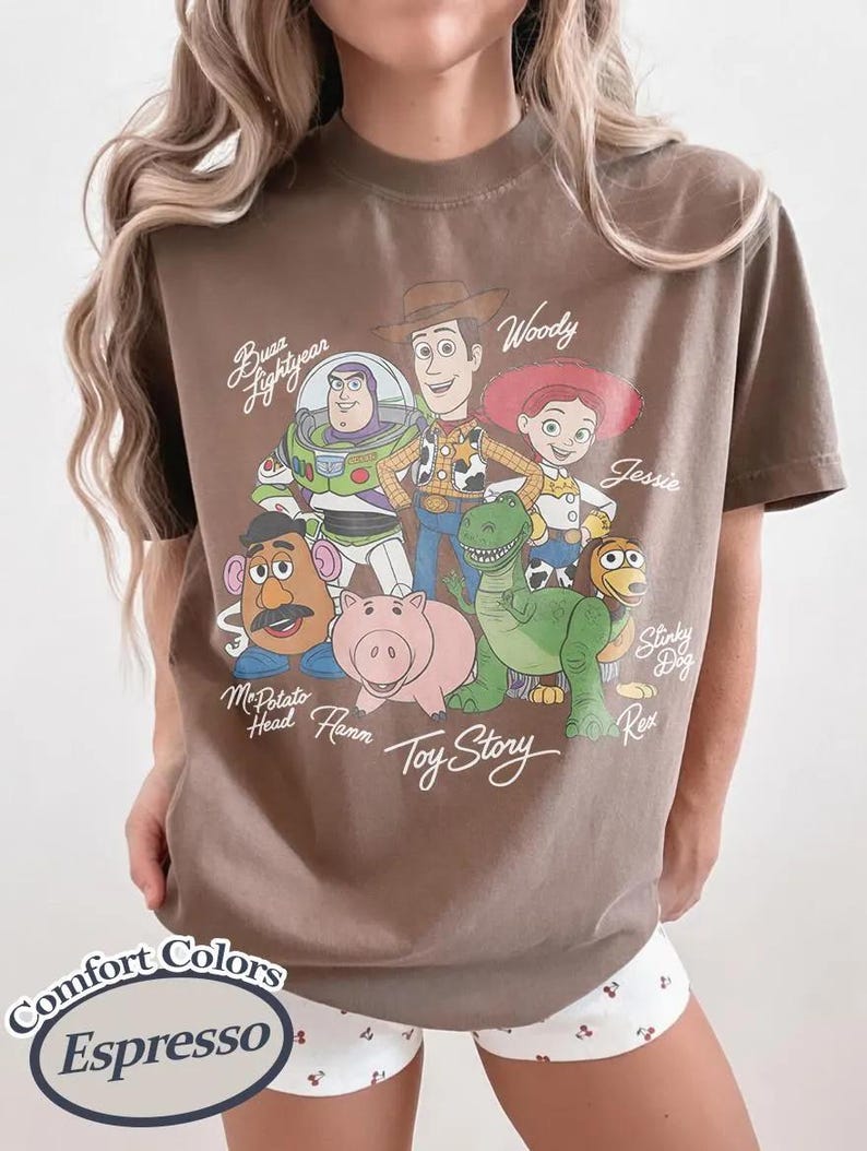 May include: Espresso-coloured t-shirt featuring a graphic of Toy Story characters including Buzz Lightyear, Woody, and Jessie. The shirt also has the text "Toy Story" and "Comfort Colors" printed on it.
