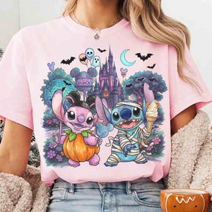 May include: A light pink t-shirt featuring a graphic print of two blue alien characters. One character is inside a pumpkin, and the other is wrapped in bandages like a mummy holding an ice cream cone. The background shows a castle and park.